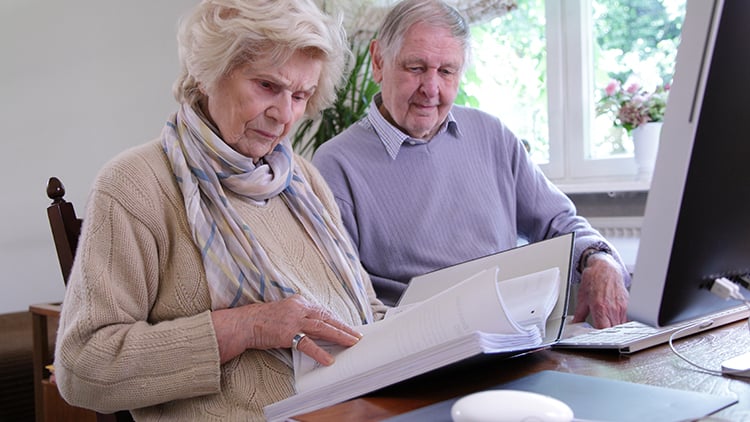  Senior couple reviewing critical documents at a table with a binder of documents and desktop computer.