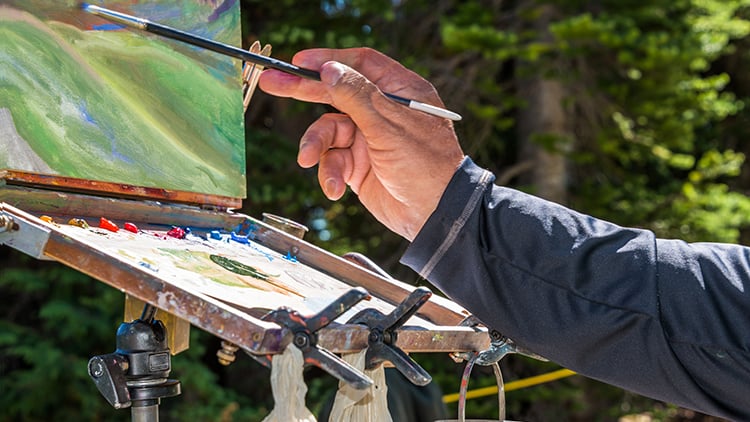 Senior man painting landscape image outdoors with oil paints